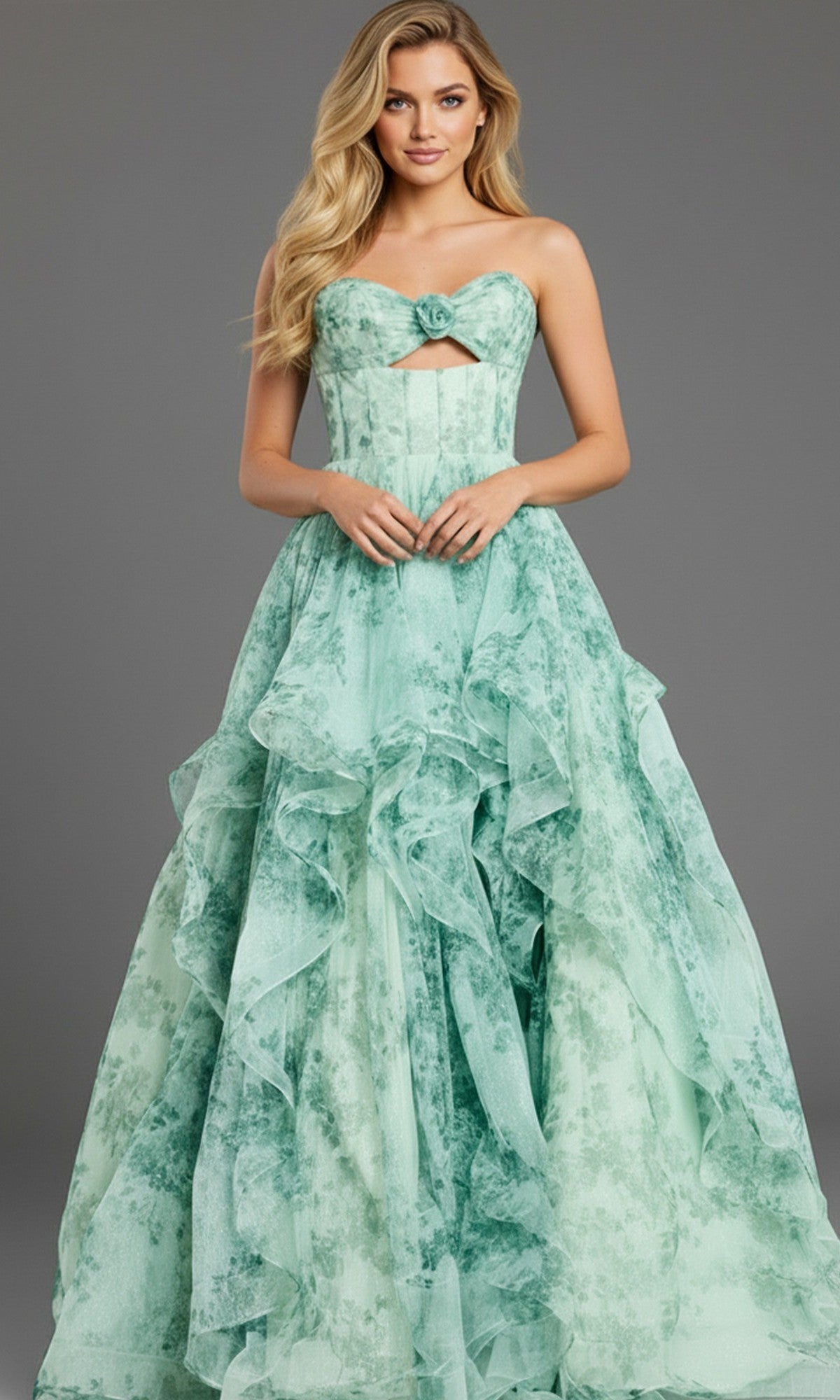 A woman wears the Strapless Green Watercolor Prom Gown: Jovani 49777, featuring floral patterns, a ruffled skirt, and a chest cutout, as she stands against a plain gray background.